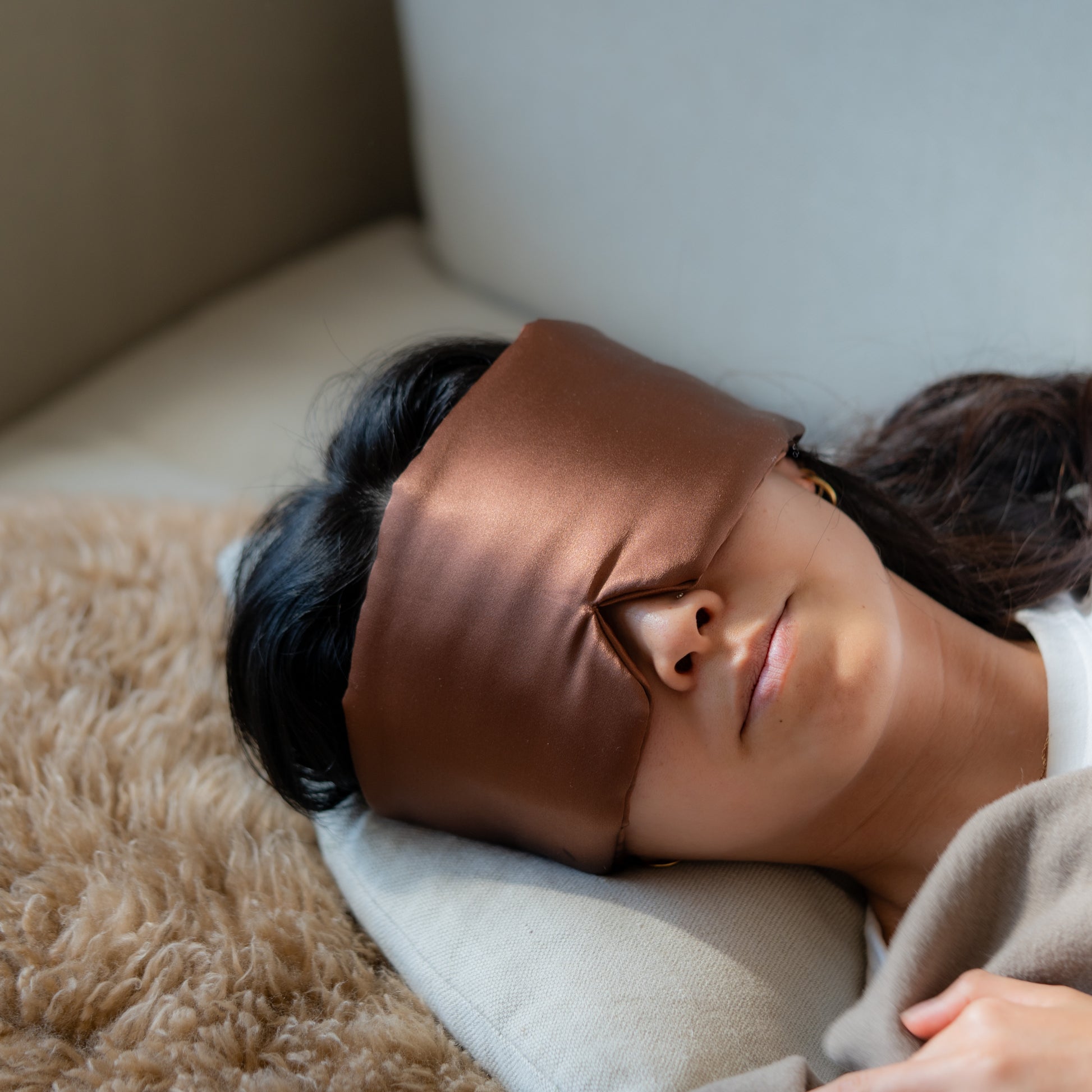 Person wearing a brown silk sleep mask lying on a pillow with a neutral wool background