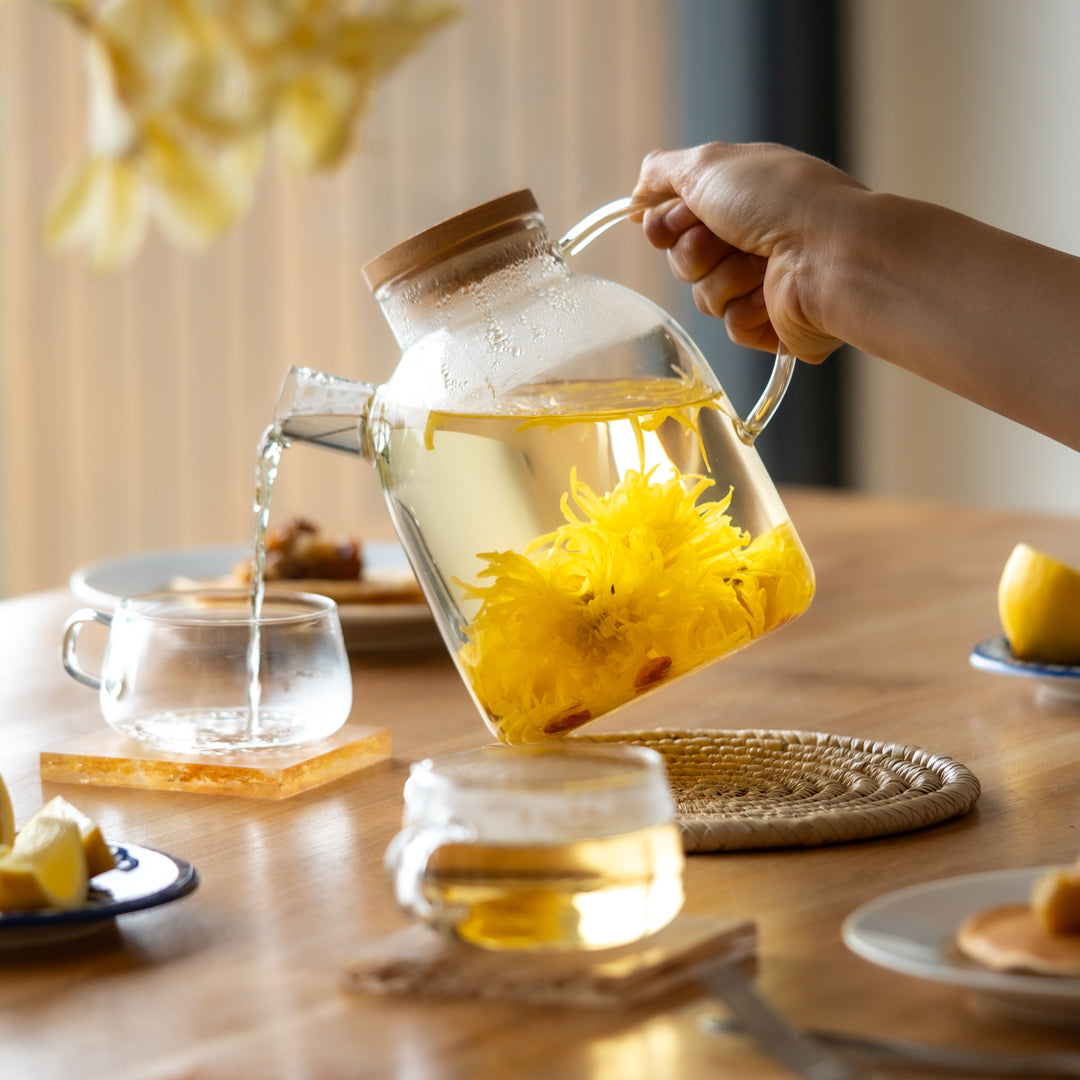 yellow chrysanthemum tea with goji berries in a glass teapot being poured into a glass mug. snacks on the table. 