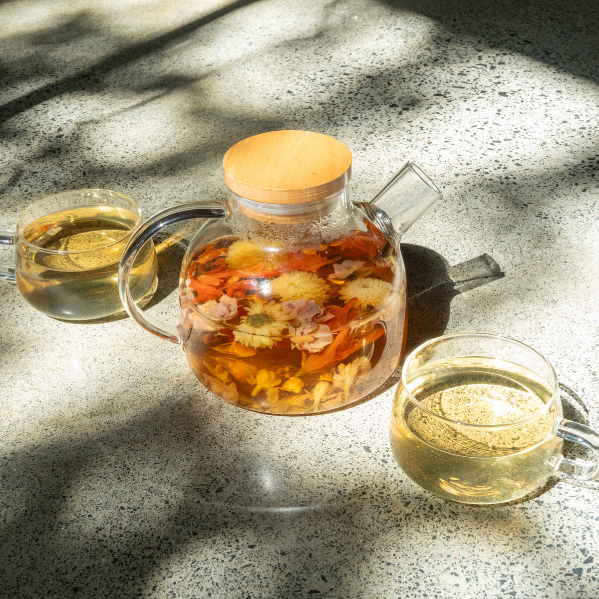 Glass teapot with inner bloom tea brewing with wooden lid and two cups on a concrete surface in natural lighting