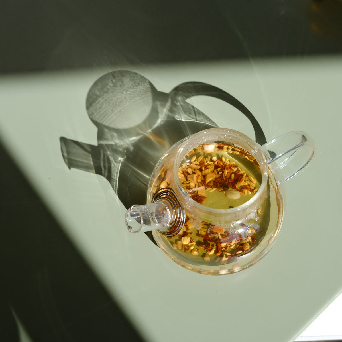 A clear glass teapot with a built-in stainless steel strainer, placed on a table with natural lighting.